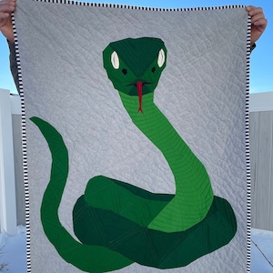 Puede incluir: Una colcha de serpiente verde con fondo blanco y borde de rayas blancas y negras. La serpiente está enroscada y tiene la lengua roja.