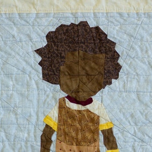 May include: A brown and yellow patchwork quilt block depicting a child wearing a brown dress with a yellow belt and yellow shoes. The child has brown hair and a brown face.