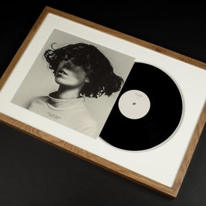 May include: A framed album cover featuring a black and white portrait of a person with dark, curly hair. A black vinyl record is partially visible. The frame is made of light-colored wood.