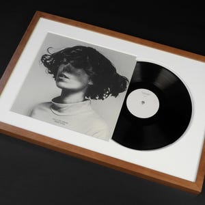 May include: A framed album cover and vinyl record. The album cover is a black and white photograph of a person with curly hair and a turtleneck. The vinyl record is black with a white label. The frame is wood.