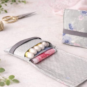 Könnte beinhalten: Eine hellgraue Stofftasche mit Zickzackmuster, geöffnet, um Wattestäbchen und gemusterten Stoff freizulegen. Eine passende geschlossene Tasche ist im Hintergrund zu sehen. Ein graues Gummiband sichert die offene Tasche.