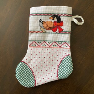 Könnte beinhalten: Weißer Weihnachtssocken mit einem grün-roten Stickmuster. Der Socken zeigt ein Kreuzstichbild eines Entchens mit einer roten Schleife.