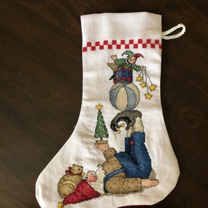 Könnte beinhalten: Weißer Weihnachtsstrumpf mit Kreuzstichmuster. Das Design zeigt einen Gaukler, der auf einem Ball balanciert, einen Weihnachtsbaum hält und den Weihnachtsmann mit einem Geschenkesack. Die Oberseite hat ein rot-weißes Karomuster.
