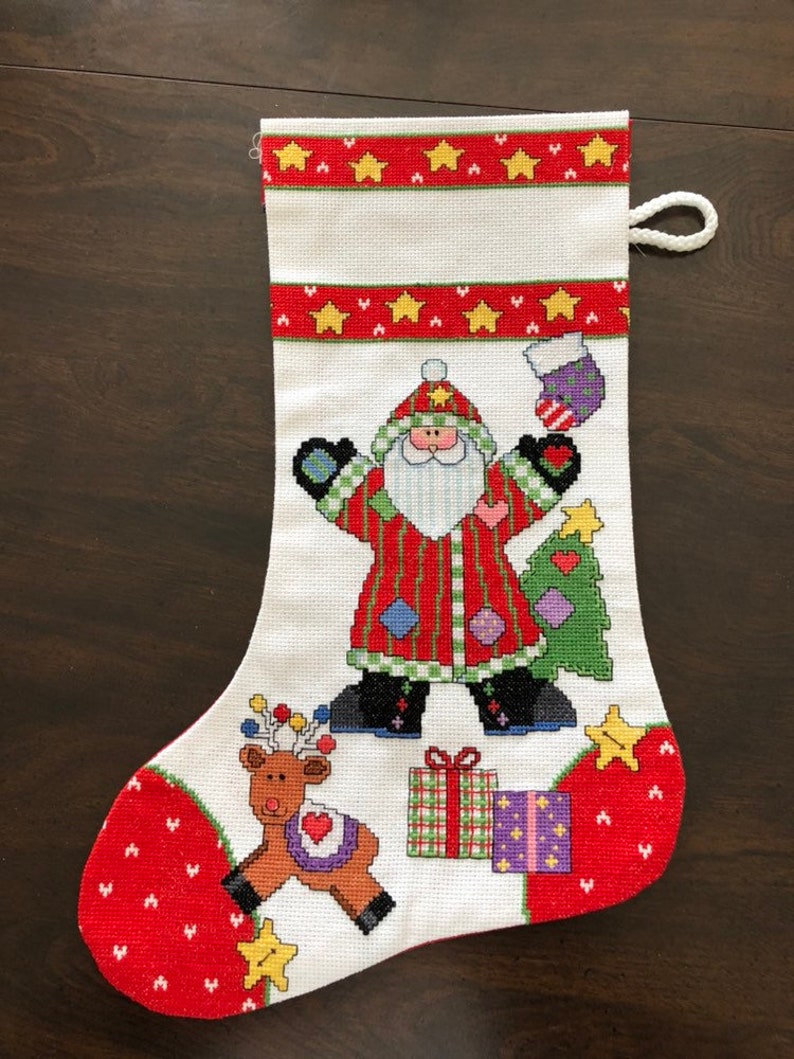 K&ouml;nnte beinhalten: Wei&szlig;er Weihnachtssocken mit rotem B&uuml;ndchen und rotem und wei&szlig;em Herzmuster. Der Socken zeigt eine gestickte Abbildung des Weihnachtsmanns, der ein Geschenk und einen Weihnachtsbaum h&auml;lt, ein Rentier und Geschenke.
