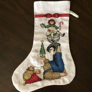 Könnte beinhalten: Ein weißer Weihnachtssocken mit einem roten Band und einem Kreuzstich-Design eines Zwergs, der einen Weihnachtsbaum hält, einer Puppe, die auf einer Kugel sitzt, und einer Katze.