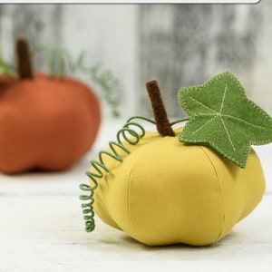 May include: Two felt pumpkins, one orange and one yellow, with green felt leaves and stems. The pumpkins are on a white wooden surface. The image includes the text "PUMPKIN SEWING PATTERN & TUTORIAL" and "PDF".