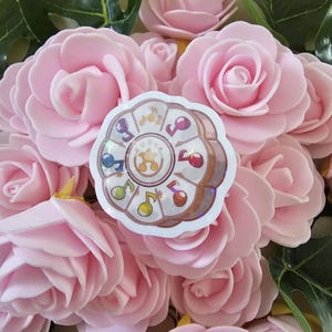 Peut inclure: Un autocollant circulaire avec des notes de musique et une étoile au centre, avec un effet holographique. Il est posé sur un fond de roses roses.