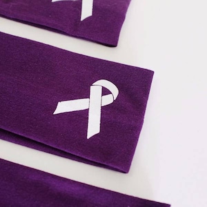 Puede incluir: Tres diademas de tela morada con un dise&ntilde;o de cinta blanca. Las diademas est&aacute;n dobladas y apiladas una encima de la otra.