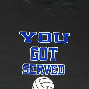 May include: Black t-shirt with the text "YOU GOT SERVED" in blue and white lettering, along with a white volleyball graphic. The shirt is a casual wear item, perfect for sports enthusiasts.