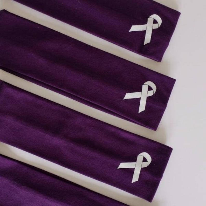 Puede incluir: Cuatro diademas de tela morada con un dise&ntilde;o de cinta blanca. Las diademas est&aacute;n apiladas una encima de la otra.