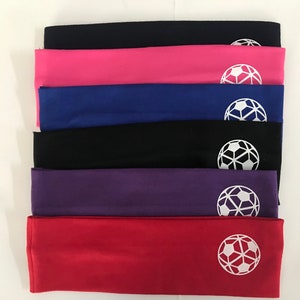 May include: A stack of six colorful headbands in black, pink, blue, black, purple, and red. Each headband has a white soccer ball graphic on the front. The headbands are made of a soft, stretchy material.