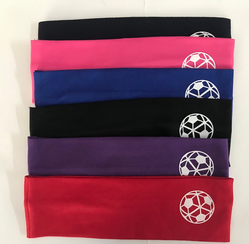 May include: A stack of six colorful headbands in black, pink, blue, black, purple, and red. Each headband has a white soccer ball graphic on the front. The headbands are made of a soft, stretchy material.