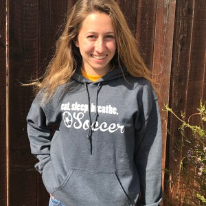 May include: A charcoal gray hooded sweatshirt with the text "eat. sleep. breathe. Soccer" in white script. The sweatshirt has a front pocket and a drawstring hood. The person is wearing blue jeans.