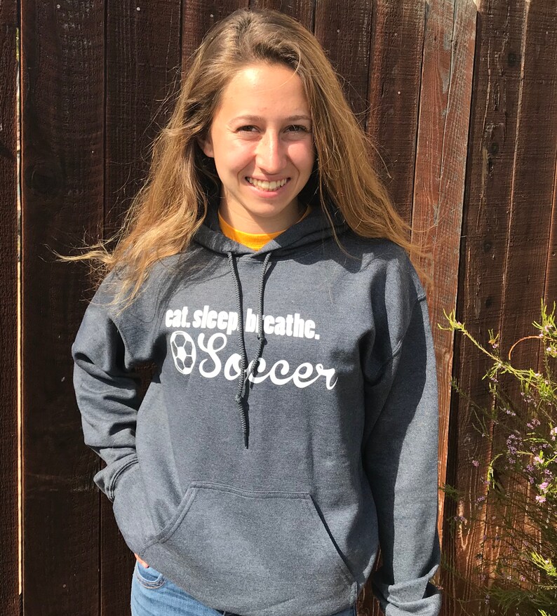 May include: A charcoal gray hooded sweatshirt with the text "eat. sleep. breathe. Soccer" in white script. The sweatshirt has a front pocket and a drawstring hood. The person is wearing blue jeans.