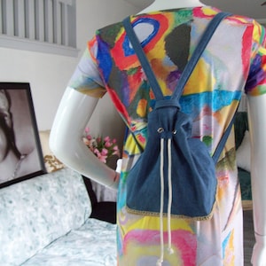 May include: A denim drawstring backpack with white drawstrings, worn over a colorful abstract print shirt.