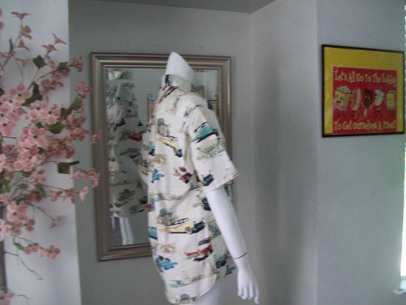 May include: A white button-down shirt with a colorful print of vintage cars. The shirt has short sleeves and a relaxed fit.