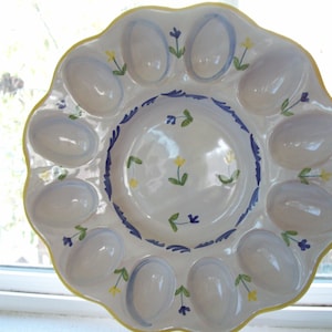 May include: White ceramic egg serving platter with a scalloped edge and a yellow rim. The platter has egg-shaped indentations and is decorated with blue and yellow floral accents. A decorative piece for serving eggs.