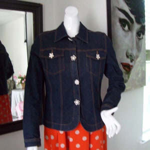 May include: A dark blue denim jacket with white flower-shaped buttons. The jacket has a classic style with a pointed collar and two front pockets. The jacket is worn over a red and white polka dot skirt.