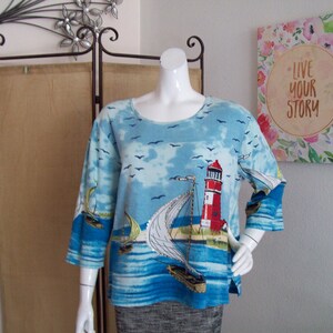 May include: A blue and white long-sleeved shirt with a lighthouse and sailboat design. The shirt has a round neckline and a relaxed fit.