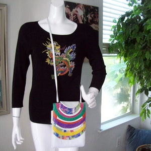 May include: White crossbody bag with a colorful, curved design on the front. The bag is worn by a mannequin wearing a black long-sleeve shirt with a dragon embroidery. The bag has a white strap.