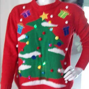 May include: Red Christmas sweater with a green Christmas tree design, white clouds, and colorful ornaments. The sweater also features small gift box designs and a yellow star on top of the tree. The sweater is displayed on a mannequin.