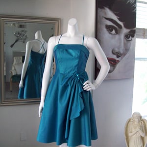 May include: Teal-colored formal dress on a mannequin. The dress features a sweetheart neckline, spaghetti straps, and a draped accent on the waist. The skirt is flared. A mirror reflects the dress's back. An Audrey Hepburn portrait hangs in the background.
