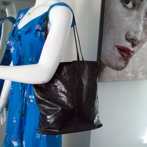 May include: A black leather tote bag with a slightly textured surface. The bag has two straps and is hanging on the arm of a mannequin.