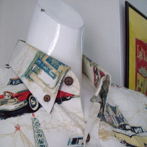 May include: A white button-down shirt with a vintage car and roadside Americana print. The shirt features a red and white car, a gas station, and a tower. The collar is folded down and the shirt is on a white mannequin.