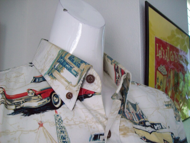 May include: A white button-down shirt with a vintage car and roadside Americana print. The shirt features a red and white car, a gas station, and a tower. The collar is folded down and the shirt is on a white mannequin.