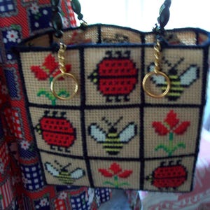 May include: A beige plastic canvas tote bag featuring a grid pattern with cross-stitched motifs of red ladybugs, yellow and black bees, and red flowers. The bag has black trim, two gold-tone metal ring handles, and black beaded accents.