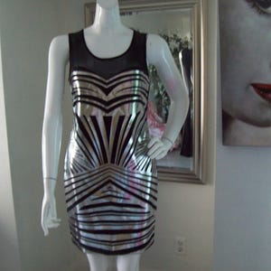 May include: A sleeveless, bodycon dress on a mannequin. The dress features a black mesh neckline and a silver and black geometric pattern. The dress is knee-length and has a fitted silhouette, suitable for a party or special occasion.