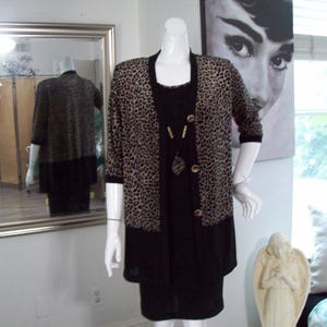 May include: A mannequin displays a black dress with a leopard print cardigan. The cardigan has a brown and black pattern and gold buttons. A long necklace with a pendant is visible. The outfit is in a room with a mirror and a portrait.