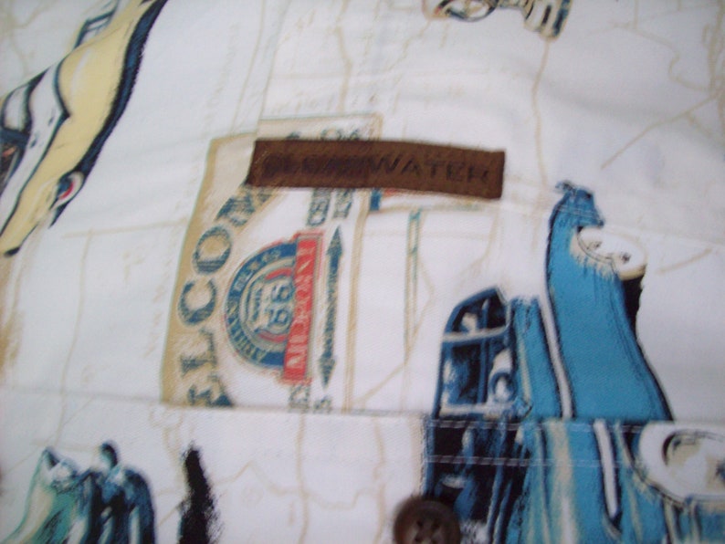 May include: White button-up shirt with a brown leather patch that says "The Navigator". The shirt has a blue and white pattern of cars and the text "El Conquistador" on the left side.
