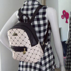 May include: A cream-colored quilted backpack with black stitching and a black top handle. The bag features a front zippered pocket with a gold-toned heart-shaped charm, black shoulder straps, and a small rectangular gold logo plate on the front.
