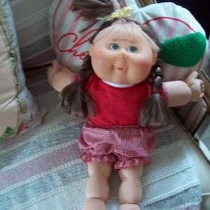 May include: A vintage Cabbage Patch Kids doll with blue eyes, wearing a red shirt and checkered shorts. The doll has brown pigtails and a yellow bow. The doll is holding a heart-shaped pillow.