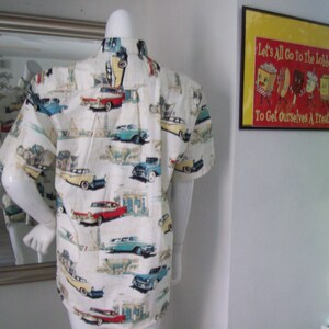 May include: A white short-sleeved button-down shirt with a vintage car print. The shirt features a variety of classic cars in different colors, including red, blue, yellow, and green. The cars are set against a white background with a subtle pattern of buildings and trees.