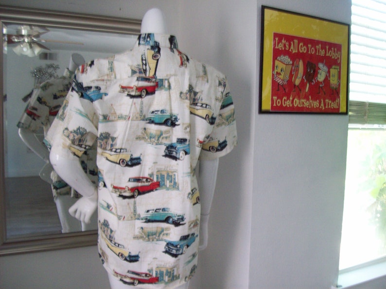 May include: A white short-sleeved button-down shirt with a vintage car print. The shirt features a variety of classic cars in different colors, including red, blue, yellow, and green. The cars are set against a white background with a subtle pattern of buildings and trees.