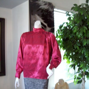 May include: A pink satin blouse with a high neck and long sleeves. The blouse has a subtle floral pattern and is displayed on a mannequin.
