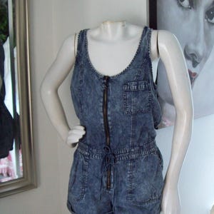 May include: A sleeveless denim romper in a light blue wash, featuring a front zipper, chest pocket, and drawstring waist. The shorts have pockets. The garment is displayed on a mannequin.