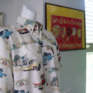 May include: A white button-down shirt with a vintage car print. The shirt has a short sleeve and a collar. The shirt is on a mannequin in front of a wall with a framed poster that says "Let's All Go To The Lobby Get Ourselves A Treat!"
