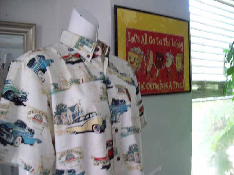 May include: A white button-down shirt with a vintage car print. The shirt has a short sleeve and a collar. The shirt is on a mannequin in front of a wall with a framed poster that says "Let's All Go To The Lobby Get Ourselves A Treat!"