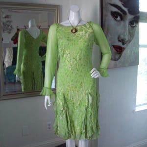 May include: A green, sheer, ruffled dress with long sleeves and a beaded necklace. The dress is on a mannequin in a room with a mirror and a painting.
