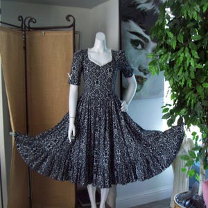 May include: A vintage-style dress on a mannequin. The dress features a black and white patterned design, short sleeves, and a square neckline with white trim. The skirt flares out, creating a full silhouette.