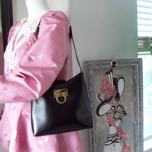 May include: A black leather shoulder bag with a gold clasp and a long strap. The bag is worn by a mannequin wearing a pink satin blouse with a white lace collar.