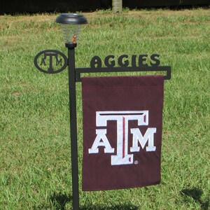 May include: A black metal garden flag stand with a maroon flag featuring a white "A&M" logo and the text "Aggies". The stand has a solar-powered light on top.