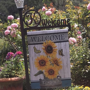 Könnte beinhalten: Dekoratives Gartenschild mit dem Wort "WELCOME" in weißen Buchstaben. Das Schild zeigt ein Sonnenblumen- und Schmetterlingsdesign auf einem Holzgrund. Montiert auf einem schwarzen Metallpfahl mit Solarlicht.