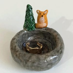 Peut inclure: Un porte-bague en céramique avec une figurine de petit renard orange et un arbre vert sur une base grise texturée. Une bague dorée repose à l'intérieur du plat. L'objet est unique et fait à la main.