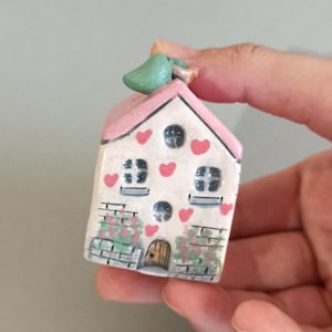 May include: A small, ceramic house figurine with a pink roof and white walls decorated with red hearts. The house has two windows, a door, and a green bird perched on top. The walls have a brick pattern with green and pink floral accents.