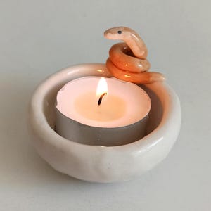 以下が含まれることがあります： 白いセラミック製のキャンドルホルダーで、上に小さな巻き付けられたヘビの置物が置かれています。ティーライトキャンドルがホルダーの中で燃えています。
