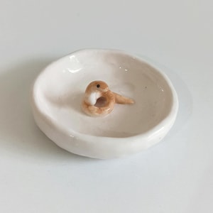 Puede incluir: Un pequeño plato de cerámica blanquecina con una figura de serpiente enrollada. La serpiente es de color beige y blanco, con un pequeño ojo oscuro. El plato tiene una forma ligeramente irregular y un acabado brillante. El diseño es minimalista y artesanal.